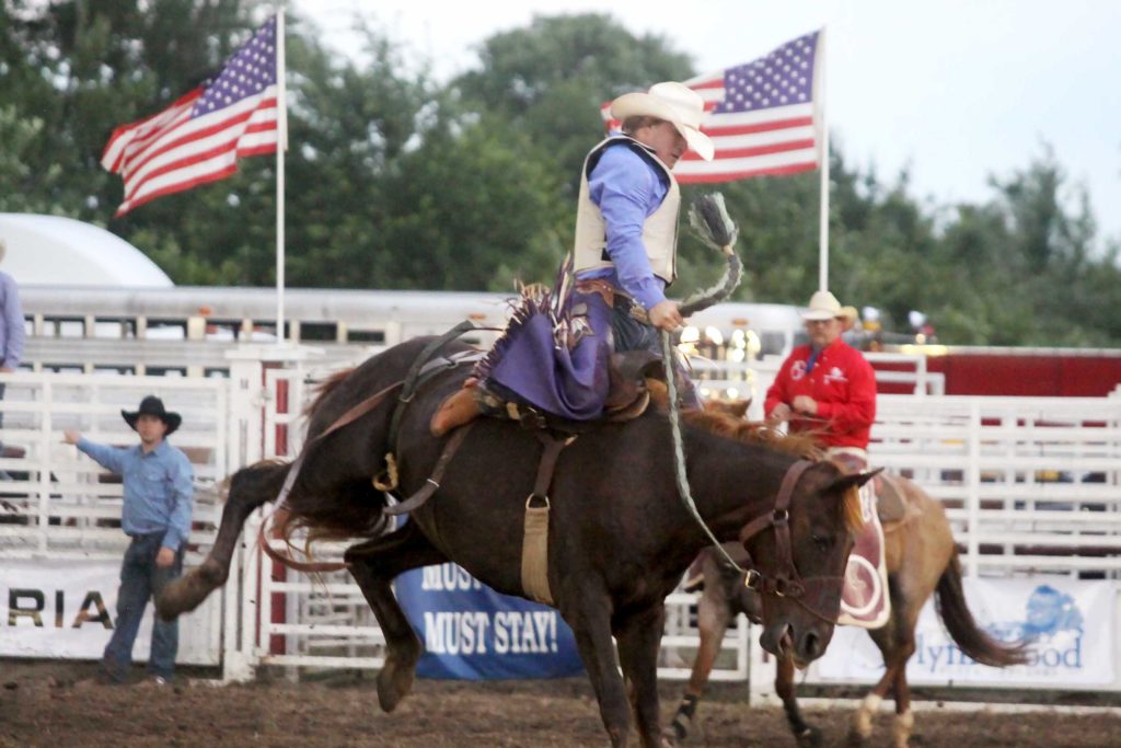 man on a riding a horse rodeo style with his arm outstretched for balance and wearing a Stetson hat american flags lining the high white fencing in the background and a man in a blue shirt on the fence along with a man in a red shirt at the fence