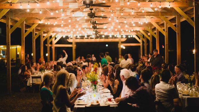 harvest dinner photograph, strung lights cover the ceiling of a barn with crowded rows of tables