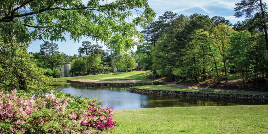 photograph of the rolling greens and a water hazard at Golden Hills Golf & Country Club. Sunny day, trees are green and to the lower left azaleas are blooming in shades of pink