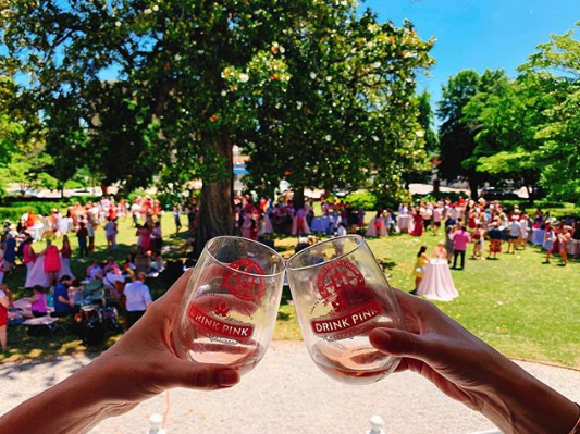 2 hands clinking wine glasses with drink pink logo on the glasses on a sunny afternoon with a crowd of people in the background