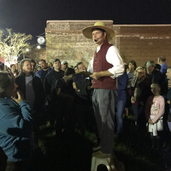 a night time photo of museum director jr fennel dressed in early american costume and hat standing above a crowd of tour goers with a headpiece microphone on