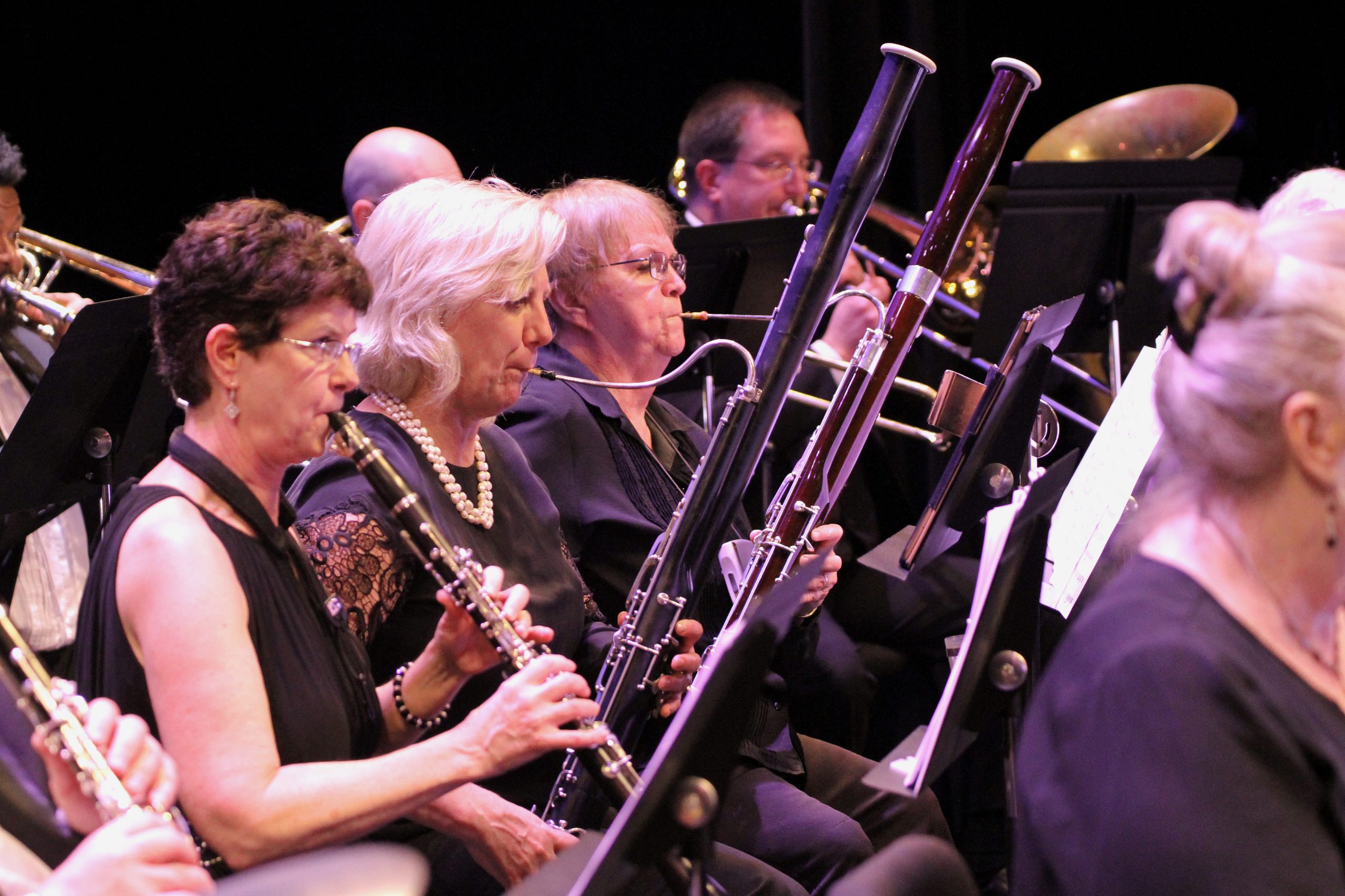 women and men seated playing playing woodwind instruments watching their music on stands in front of them