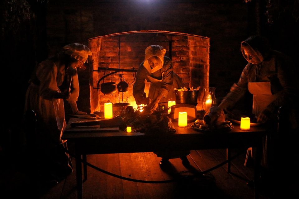 historic reinactors recreating a nighttime kitchen scene with food and candles on the table and women working on dinner