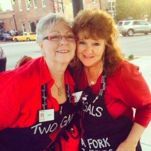 janet and gloria of two gals and a fork leaning into each other smiling at the camera wearing red blouses and black printed aprons