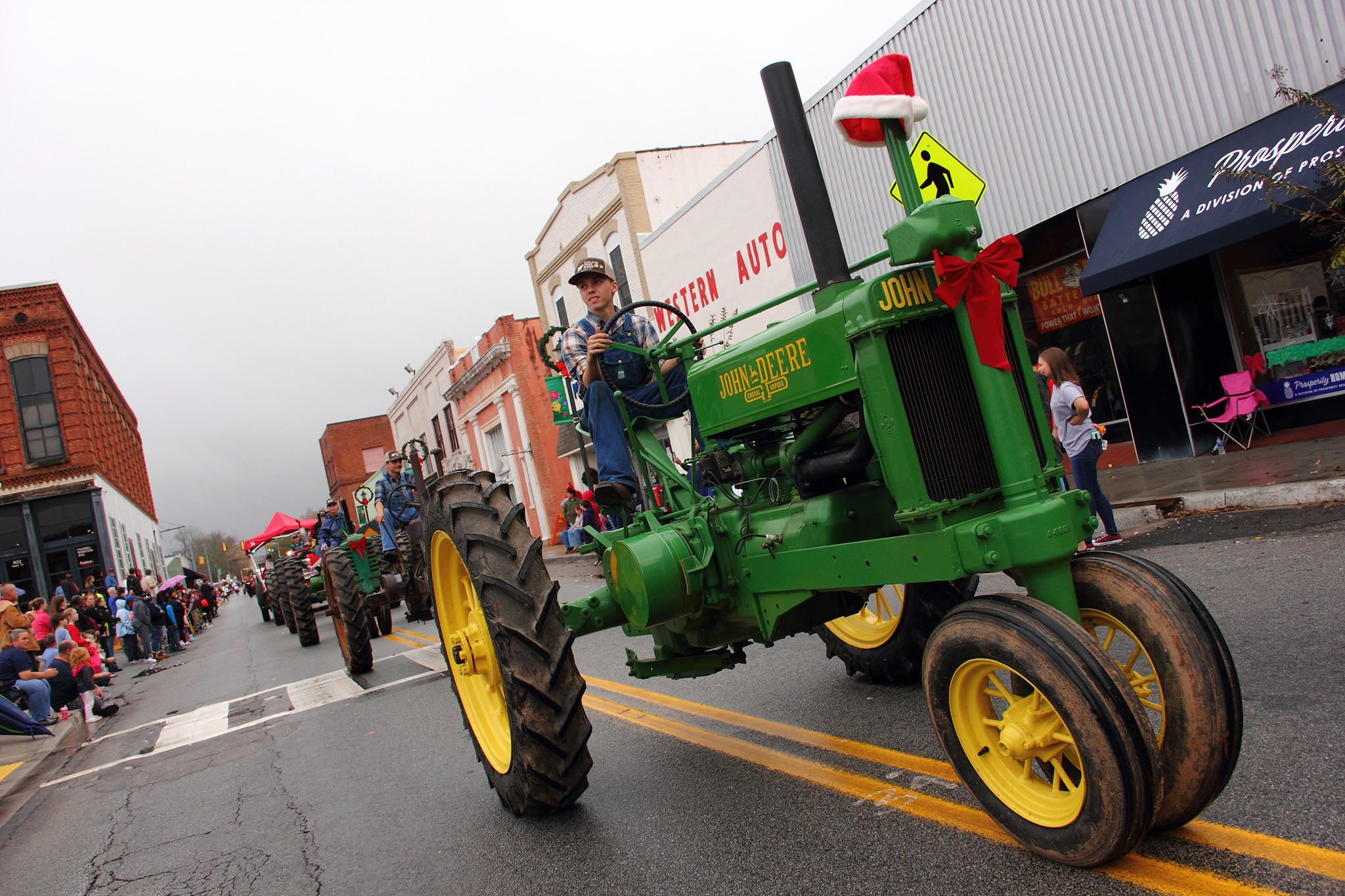 young man dressed as a farmer on an antique john deere tractor on a rainy day in small town in them middle of main street for a parade of tractors behind him