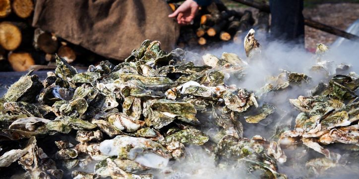 steaming oysters on a table with an apron wearing person standing in the back, only their apron and hand are visible