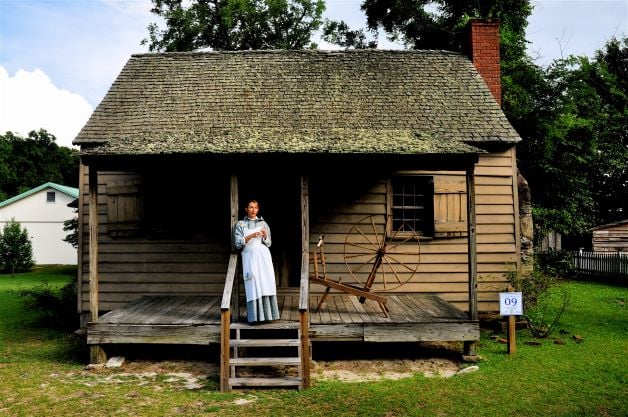 A woman in colonial period costume stands on the porch of a historic cabin on the property of the Lexington County Museum