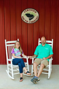 Craig and Meredith Amick sitting in white rocking chairs holding hands at their distillery beneath an SC Certified sign