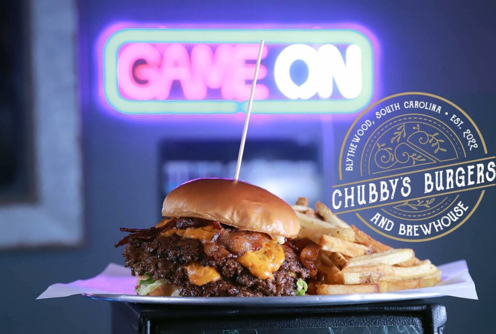 smash burger with cheese and trimmings sandwiched between a bun with a tooth pick holding it together sits next to a crispy pile of french fries on paper. The chubby's logo is present and a neon sign in the background says game on