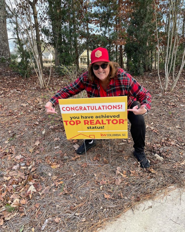 woman wearing red plaid shirt with baseball hat and sunglasses crouches and points at yellow yard sign that says top realtor
