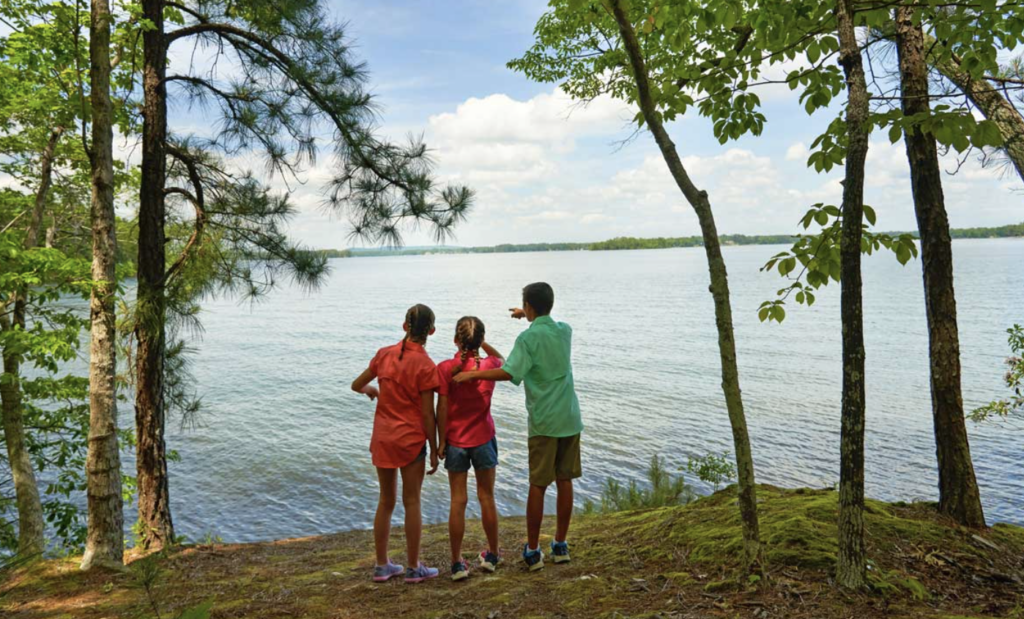 Three children standing on the shore of Lake Murray at Dreher Island State Park, surrounded by Trees.