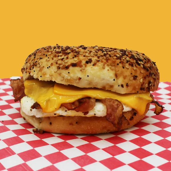 An everything bagel with bacon, egg, and american cheese on a red checkered tablecloth and a yellow background