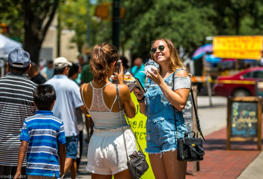 Two young women stand in the street taking a selfie, one holds a camera while the other poses with a drink. In the background people are walking down the street