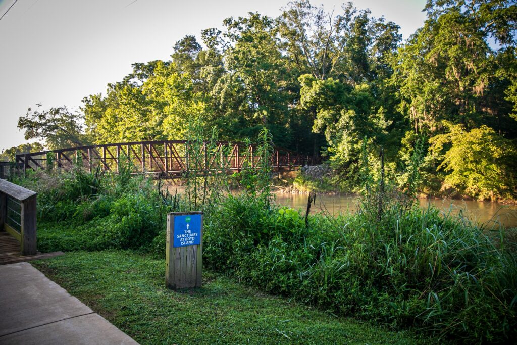 A bridge connecting the riverwalk to the Sactuary at Boyd Island crossing over the river and surrounded by greenery and trees.