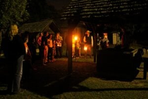 Visitors gather around a well outside the Lexington County Museum at night, only lit by glowing orange lanterns as museum staff are dressed up and sharing tales of haunted regional history.